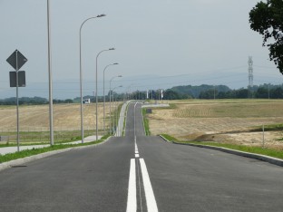 Zdjęcie przedstawia teren inwestycyjny do którego prowadzi droga publiczna. wzdłuż drogi ustawione są lampy oraz znaki drogowe. 